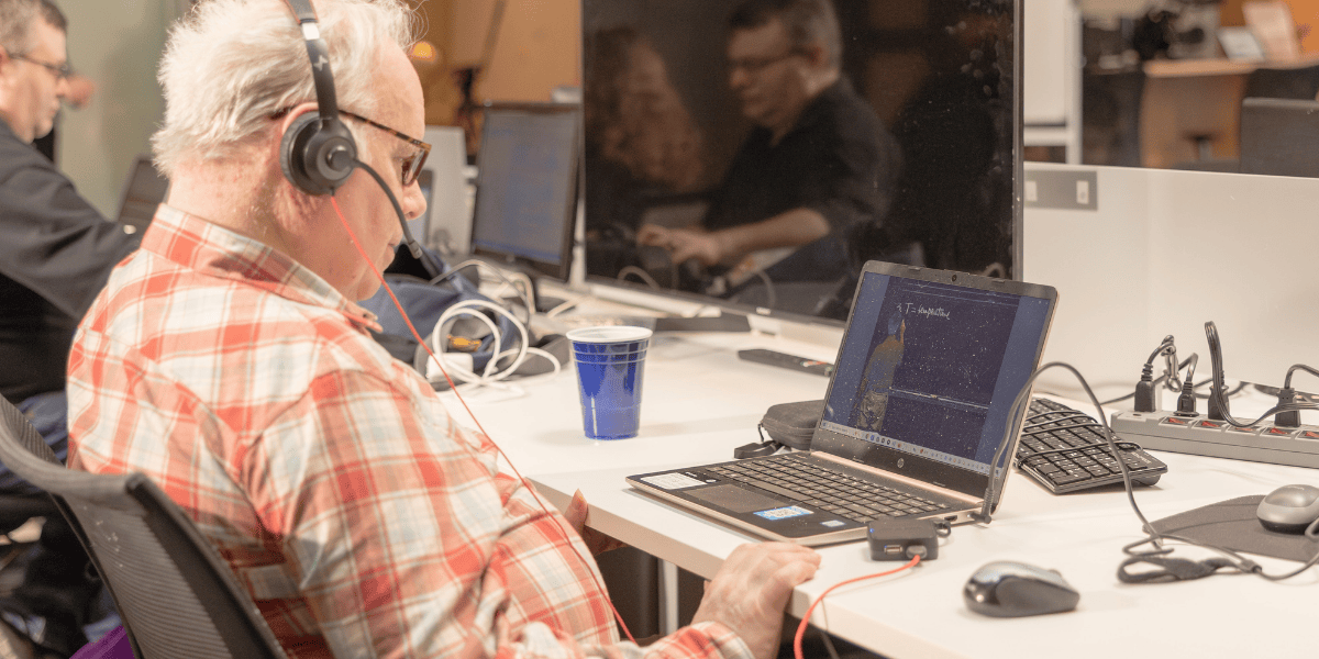 A man in his late 60s in a red plaid shirt wears headphones while working at a laptop in an open office. Two coworkers nearby are reflected in a monitor screen on the same desk.