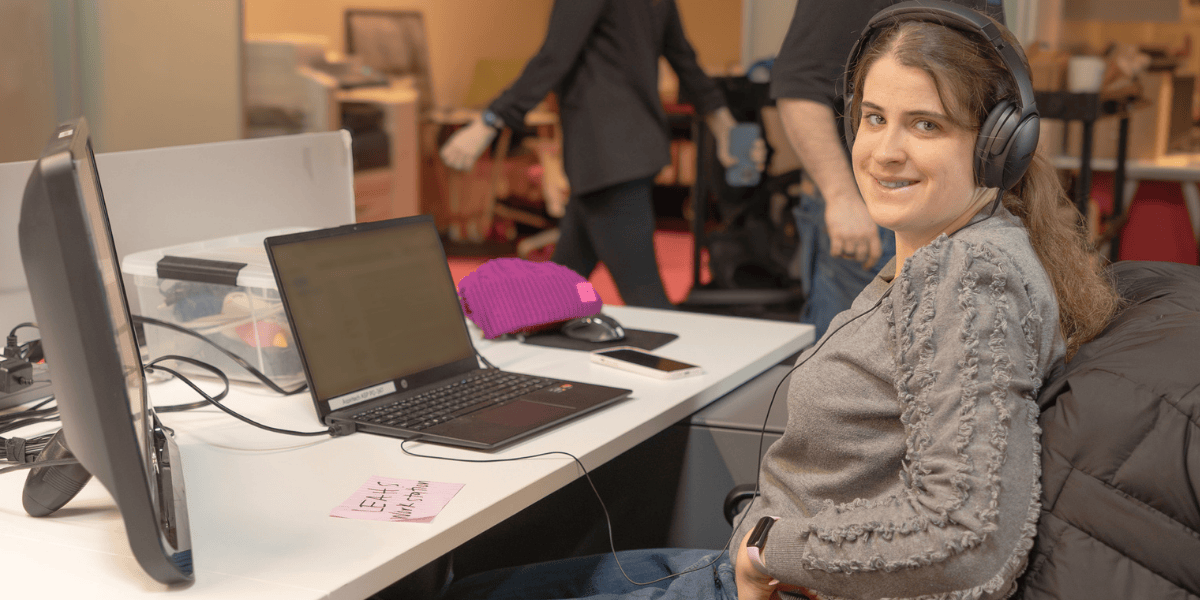 A woman with brown hair and a gray sweater smiles at the camera while wearing headphones and sitting in front of her laptop. Two other office workers are walking by in the background.